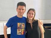 i-2 - Иван Дончев и Биляна Динкова - прави един до друг и усмихнати, в светла музикална репетиционна зала или кабинет. В центъра на кадъра мъжът е облечен в синя тениска с щампа от четири ретро оперни плаката, сред които личи надписът „Turandot“, и светли панталони. От дясната му страна стои младата жена с дълга светла коса, облечена в къса черна рокля без ръкави, като мъжът я е прегърнал приятелски през рамото. Непосредствено зад тях се вижда голям черен роял „Yamaha“, върху чийто отворен капак има разтворена нотна тетрадка със спирала, а вляво върху него лежи жълта папка. На задната бяла стена, точно над рояла, е монтиран голям правоъгълен звукоизолиращ акустичен панел със сива релефна текстура на пирамидки. Подът на помещението е покрит със светли квадратни плочки, подредени по диагонал, а в долния десен ъгъл се забелязва малка част от син стол.